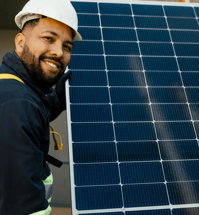 A worker wearing safety gear holds a solar panel